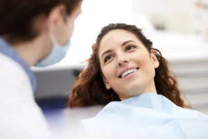 A woman with long curly hair smiles while reclining in a dental chair, looking at a blurred dentist in a blue mask, conveying comfort and care.