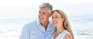 A smiling older couple stands closely on a sunny beach. The man wears a light blue shirt, while the woman is in a white sleeveless dress. The ocean waves sparkle in the background.