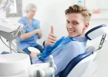 Young man in a dental chair gives a thumbs-up and smiles confidently. Dentist in the background.
