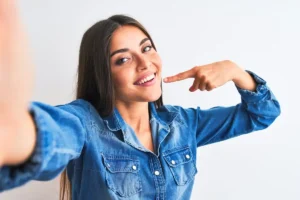 Smiling woman with long dark hair in a bright green sweater points to her cheek. She stands against a solid purple background, conveying a cheerful mood.