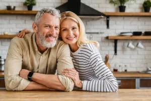 A smiling couple leans on a wooden kitchen counter, embracing. The man wears a beige shirt and smartwatch; the woman wears a striped shirt. Cozy ambiance.