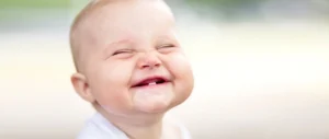 A smiling baby with eyes closed and a partially visible tooth grins joyfully against a blurred background, conveying pure happiness and innocence.