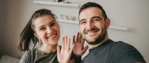 A smiling couple waves at the camera indoors, exuding happiness and warmth. They stand close together, framed by a blurred background with shelves.