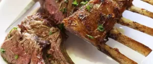 Grilled rack of lamb on a white plate, featuring browned, seasoned ribs garnished with herbs, conveying a savory, appetizing presentation.