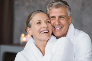 An older couple in white robes smiles warmly, embracing each other. Soft lighting and blurred candles in the background create a cozy, intimate atmosphere.