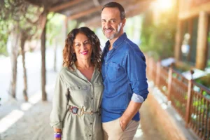 A couple smiles warmly in a sunlit outdoor corridor. The woman wears a green dress and the man a blue shirt, exuding a relaxed and happy vibe.