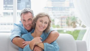A smiling couple in blue shirts embrace on a couch, with an urban cityscape and greenery visible through a sunlit window, conveying warmth and contentment.