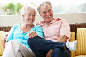 Elderly couple smiling and sitting on a patio sofa. The woman in a blue sweater rests her hand on the man's knee.