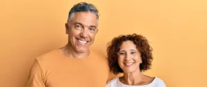 Smiling man and woman standing together against an orange background. The man wears a matching shirt; the woman wears a white top. Warm, cheerful vibe.