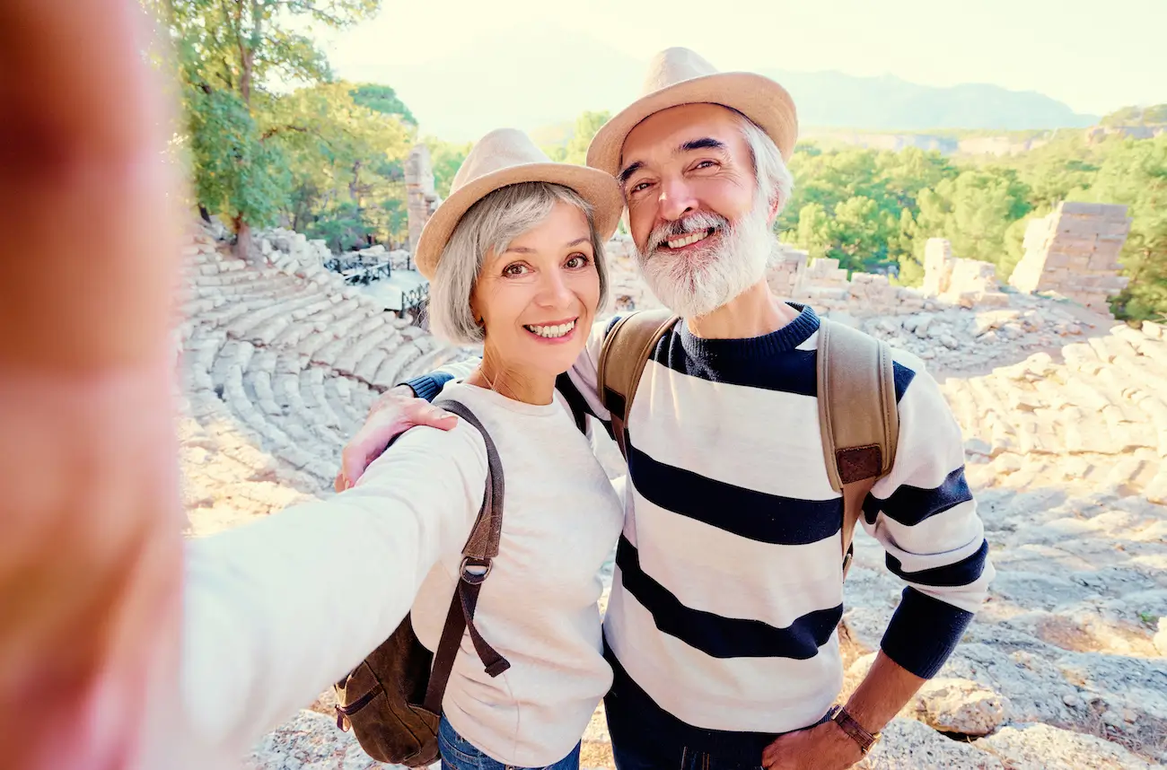 Elderly couple wearing hats and backpacks takes a cheerful selfie at an ancient amphitheater. Sunlit stone seats and green trees form the background.