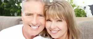Smiling older couple sitting closely on an outdoor couch, surrounded by greenery and white flowers, conveying warmth and happiness.