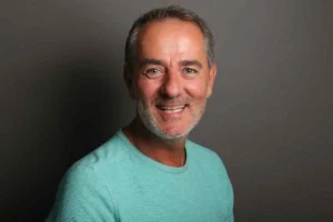A smiling man in a turquoise shirt against a gray background, exuding warmth and friendliness. His short hair and subtle beard add a casual touch.