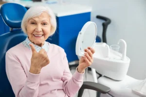 Elderly woman in a dental chair holds a mirror, smiling and giving a thumbs-up. She wears a pink sweater, conveying satisfaction and joy at a dental clinic.