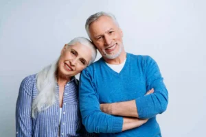 Elderly couple smiling warmly, woman with long white hair in a striped shirt leans on a man in a blue sweater, both exuding happiness and contentment.