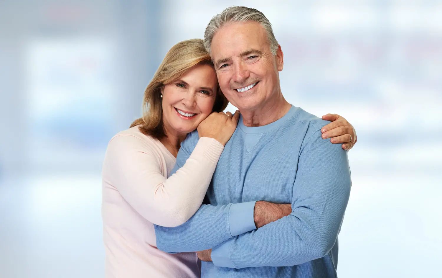 An older couple smiles warmly, embracing each other. The woman, wearing a pink sweater, leans on the man's shoulder, who wears a blue sweater. The background is softly blurred.
