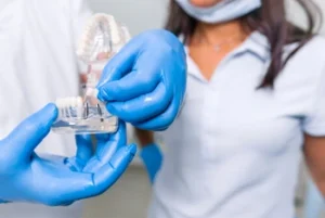 Dental professional with blue gloves holds a dental implant model while explaining to a patient wearing a mask and white shirt, conveying a clinical tone.