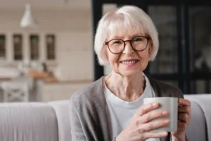 Elderly woman with short white hair and glasses smiling warmly, holding a mug in a cozy living room. The atmosphere is calm and inviting.