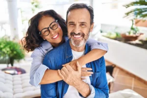 A smiling woman with glasses hugs a man from behind. They're outdoors with plants in the background, conveying warmth and happiness.