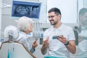 A dentist shows a senior woman a set of dental veneers in a modern clinic. They are discussing options, both smiling. An X-ray is visible on a screen nearby.