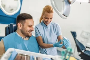 A smiling dentist shows a dental mold to a male patient seated in a dental chair. The clinic has a bright and friendly atmosphere.