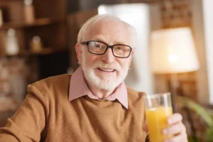Elderly man with glasses and white beard, smiling warmly while holding a glass of orange juice indoors.