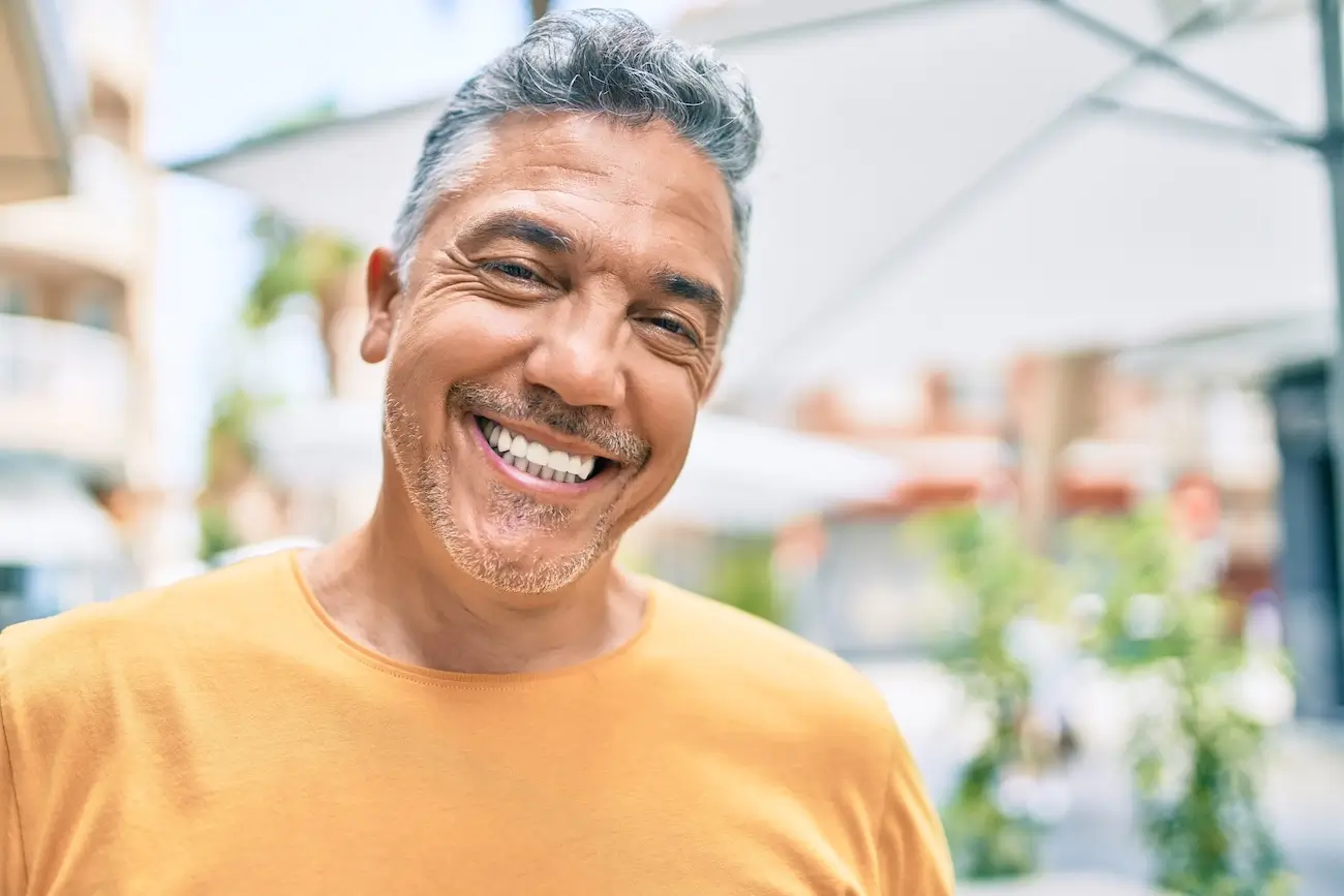 Smiling man in an orange shirt stands outdoors under a white umbrella. The bright, sunny setting and his cheerful expression convey warmth and joy.