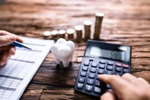 A person calculates expenses on a table with stacks of coins, a dental model, a calculator displaying 54800, and a pen over a financial document.