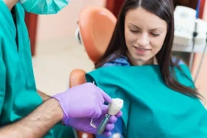 A dentist in green scrubs and purple gloves shows a dental implant model to a seated, smiling woman in a dental chair. The atmosphere is calm and informative.