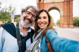 A smiling couple takes a selfie in front of a large, historic brick arch. The sky is clear, adding a joyful and relaxed atmosphere to the scene.