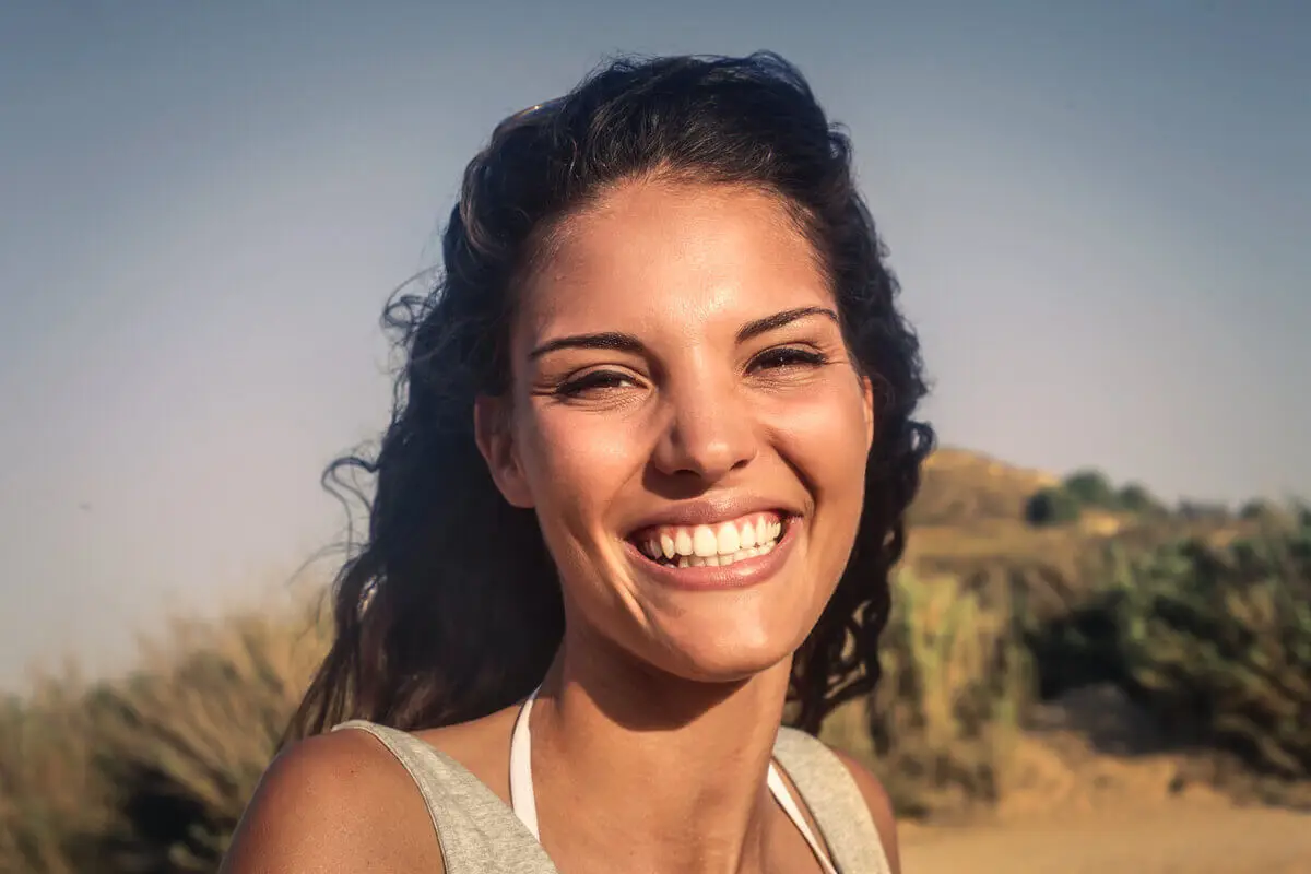 A woman with long, brown hair smiles brightly at the camera. The background is a sunny outdoor setting with blurred greenery and a clear blue sky.