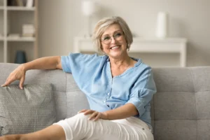 Smiling older woman with short gray hair and glasses, wearing a blue shirt and white pants, relaxing on a gray sofa in a cozy living room.