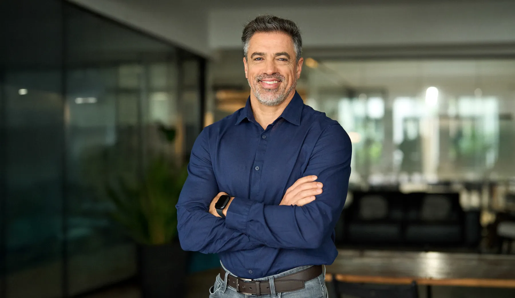 Smiling man with crossed arms in a blue shirt stands confidently in a modern office space with glass walls and a blurred background.