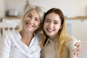 A smiling older woman with gray hair and a younger girl with long hair sit closely together, arms around each other, in a bright, cozy room.