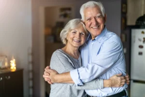An elderly couple lovingly embraces and smiles in a cozy, warmly-lit kitchen. The scene conveys a sense of happiness and togetherness.