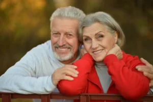 An elderly couple smiling warmly, leaning on a wooden fence. The man wears a light hoodie, and the woman a red jacket. Background shows soft-focus autumn trees.