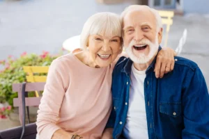 Elderly couple smiling warmly, woman in a pink sweater, man in a denim shirt, seated on a bench with flowers in the background, portraying joy.