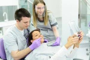 A woman in a dental chair smiles while holding a mirror. A dentist and assistant, both in gray scrubs and gloves, assist her. The setting is bright and clinical.