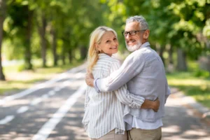An older couple smiling warmly at each other while walking in a lush park. The scene is bright and cheerful, conveying happiness and togetherness.