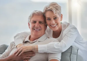 Elderly couple sitting closely on a sofa, smiling warmly at the camera. The woman gently embraces the man, conveying a sense of love and contentment.