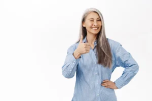Smiling woman with long gray hair gives a thumbs up, wearing a blue striped shirt. She exudes positivity and confidence against a white background.