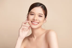 Smiling woman with short brown hair, looking at the camera. She is touching her face with one hand against a neutral beige background.