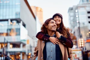 A joyful couple embraces amid an urban setting. The woman, wearing a hat, smiles brightly from behind as the man looks ahead, exuding happiness.