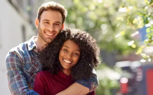 A smiling couple embraces outdoors on a sunny day. The man, wearing a plaid shirt, stands behind the woman, who has curly hair and wears a red top.