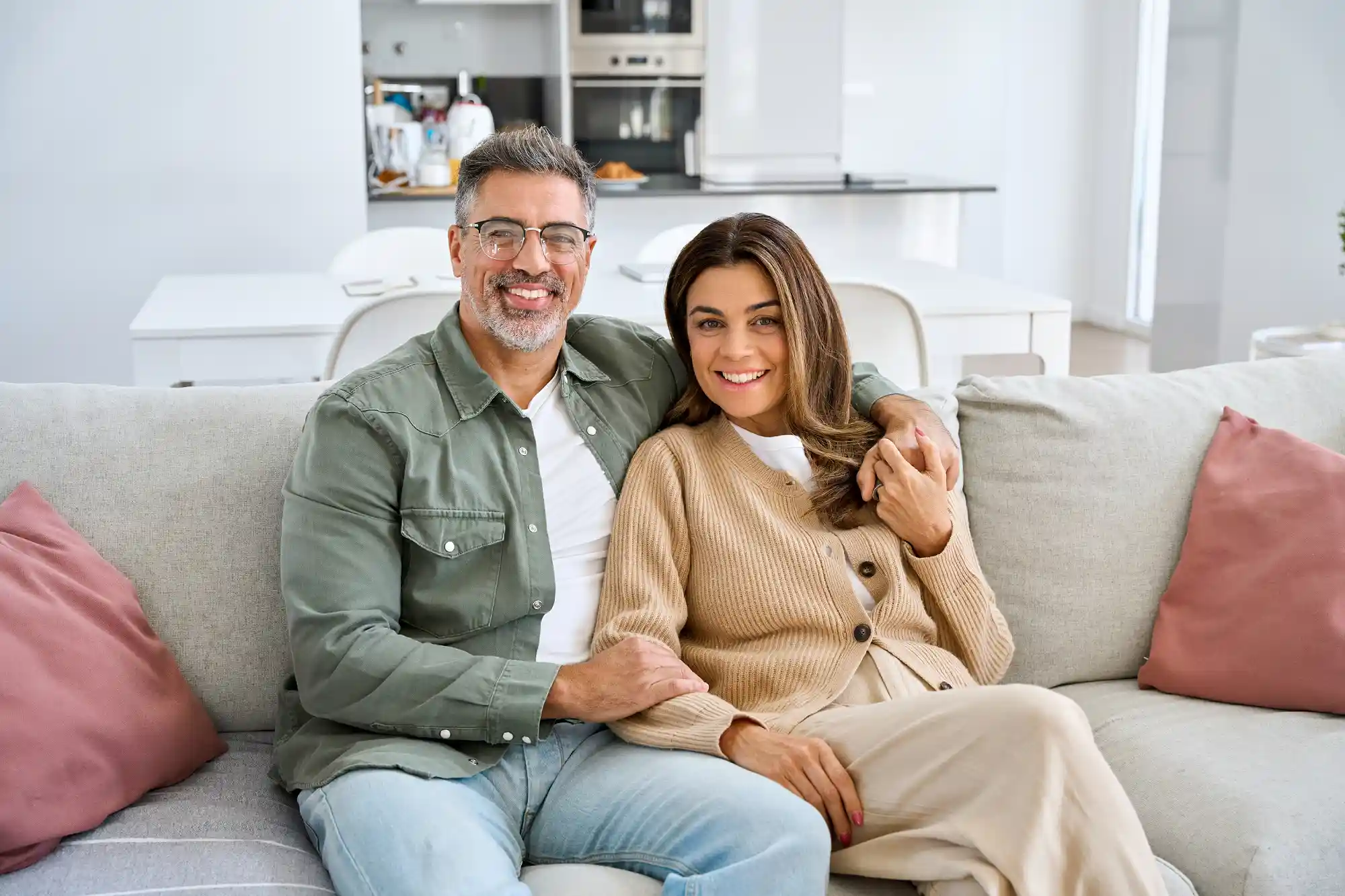 A smiling couple sits on a beige sofa in a modern living room. They are casually dressed, exuding warmth and comfort. The room is bright and airy.