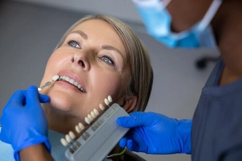 Woman at dentist's office having teeth color matched with a shade guide. Dentist wearing blue gloves and mask, performing the procedure.