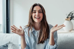 A smiling woman with long brown hair in a striped shirt sits on a couch, gesturing with open hands. A white vase with flowers is in the blurred background.