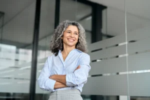 Smiling woman with curly hair, wearing a striped shirt, stands confidently with arms crossed in a modern office with glass walls.
