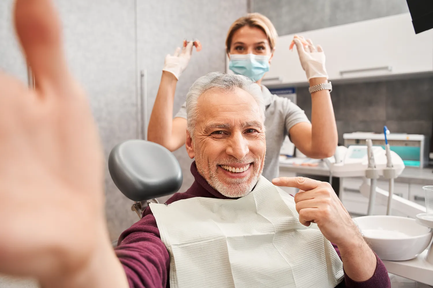 Elderly man smiling in a dental chair, pointing at his teeth. A dentist in gloves stands behind him, also smiling. A friendly, reassuring scene.
