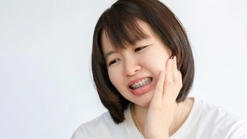 A person with shoulder-length hair and braces is holding their cheek, looking uncomfortable, possibly due to dental pain. The background is plain white.