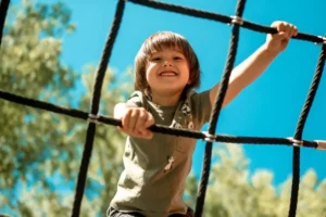 Smiling child on a rope climbing structure against a bright blue sky, with green trees in the background. The mood is joyful and adventurous.